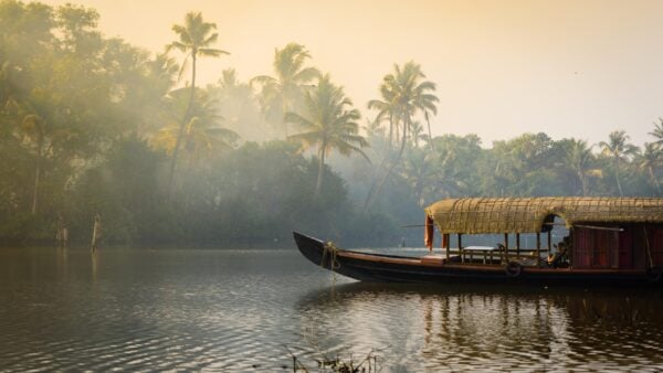 A traditional house boat is anchored on the shores of a fishing lake lined with palm trees on a misty morning in Kerala's Backwaters, India.