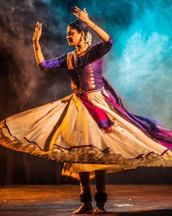 A beautiful kathak dancer in traditional dress with a a big, voluminous skirt, twirls against a smoky backdrop on stage