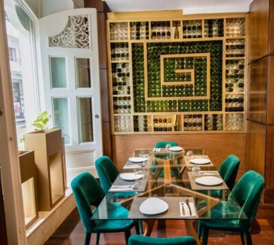 Dining area with large glass table set with plates and cutlery, with plush green seats and spiral artwork created by bottles on the wall