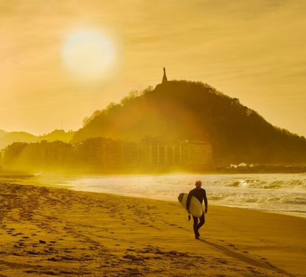 A surfer walking on the Zurriola Beach at sunset with the Monte Urgull in the background. San Sebastian, Basque Country, Guipuzcoa. Spain.