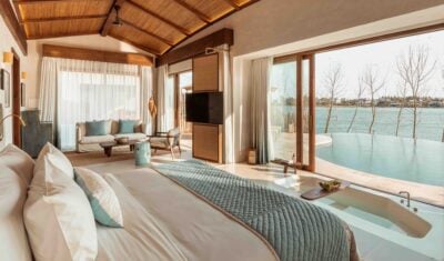 Bedroom in a villa with large glass doors open to reveal a private pool. In the room there is a large bed with cream and pale blue bedding, a sofa and a chair. At the foot of the bed there is a bath sunken into the floor