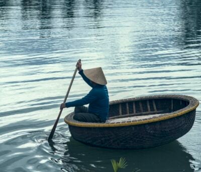 A person wearing a traditional conical hat paddling a round boat through still water