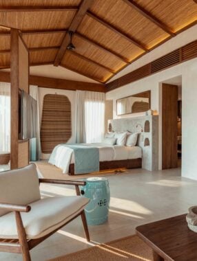 Interior bedroom of a villa with high wooden ceiling, large double bed with white bedding, and an armchair