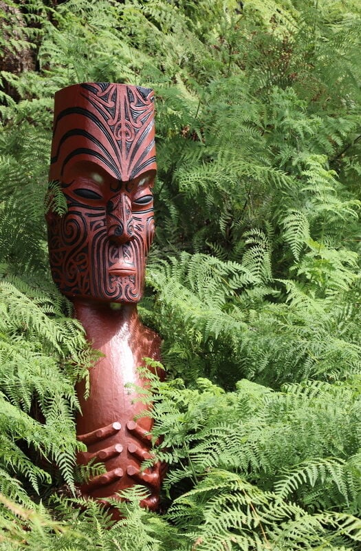 Sculpture of a Māori Totem, a carved wooden figure amongst thick fern foliage