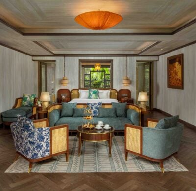 Hotel bedroom with wood panelled walls, a large double bed with white bedding, and a seating area with moss green coloured sofa and chairs. There is a small round table in the middle of the seats, laid with a teapot, two tea cups, and a cake stand.