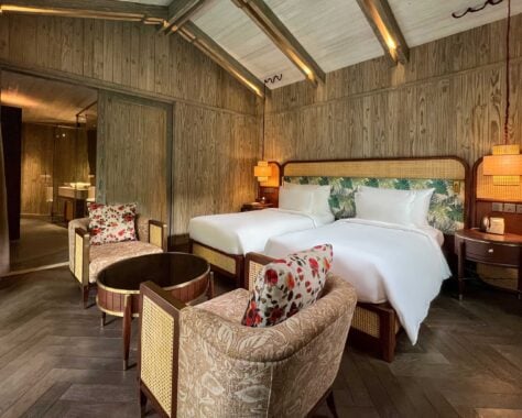 Interior view of hotel bedroom with two single beds with white bedding against a leaf-print headboard. At the end of the beds there is a small round table and two armchairs with flower-print cushions