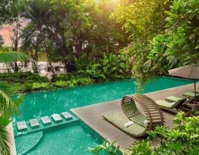 Exterior view of swimming pool with wooden decking area and sun loungers surrounded by tropical foliage