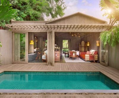 Exterior shot of a poolside villa, showing through open doors a living area with large sofa and chairs, and a bathroom with free standing tub