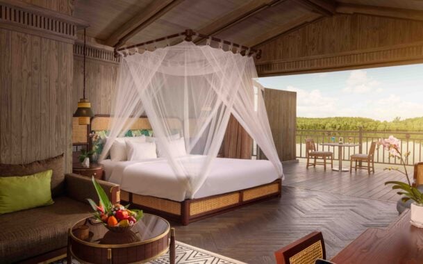 Interior view of a large bedroom with four poster bed with white bedding and net drapes, a small round table with a fruit bowl, and a large outdoor verandah with two chairs and a small table by the water