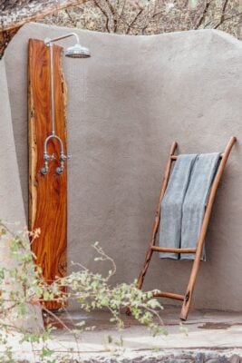 Exterior image of a hotel outdoor shower, with smooth rock privacy wall, and a large shower head mounted on a wooden panel. There is a wooden frame towel rail leaning against the wall, with two blue towels draped over it