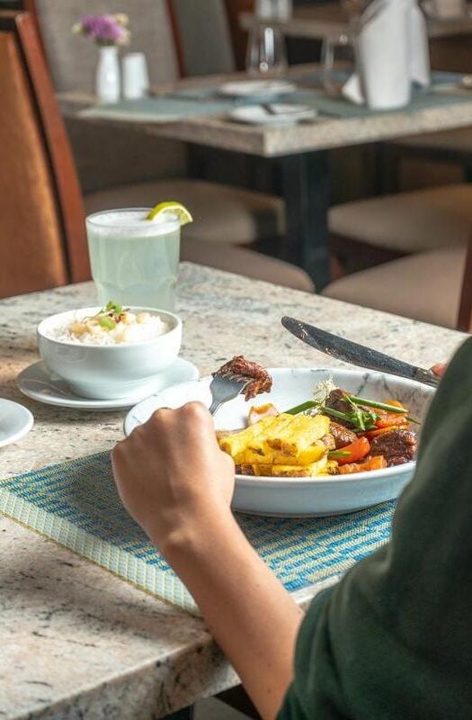 The hands of a person using a knife and fork to eat Lomo saltado out of a white bowl, with a glass of juice and a bowl of arrox con leche in the background