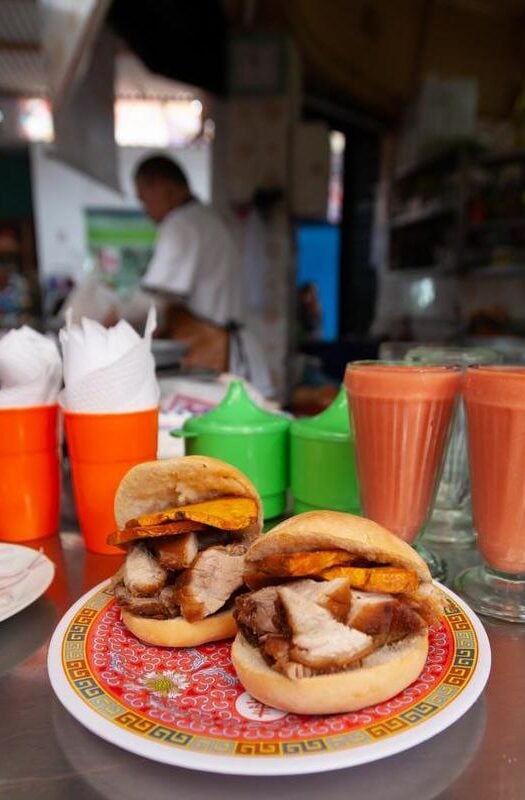 Peruvian pork sandwich on a colourful plate with glasses of orange-coloured juice and a man working in the background