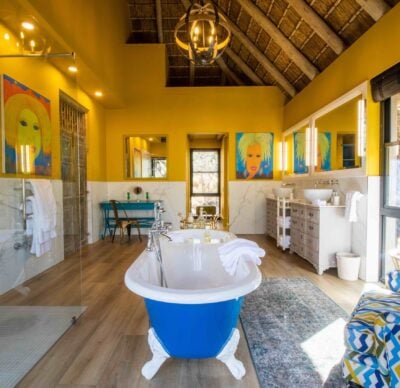 A bathroom painted in sunny yellow with a free standing blue and white tub, two sinks, and a glass-sided shower