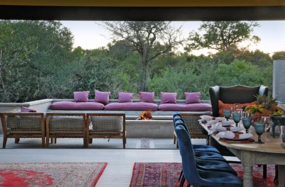 Outdoor seating area with long couch covered in pink cushions. There are also low chairs around a fire pit