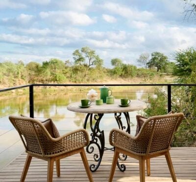 A small circular table and two wooden chairs set for tea on a wooden deck overlooking a river