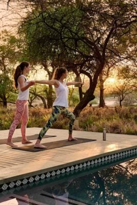 Two women practising yoga on a wooden deck by a pool at sunrise