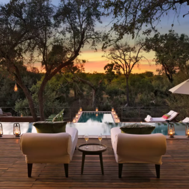 Outdoor seating area with a long infinity edge pool surrounded by lanterns, and with two sun loungers facing the pool and forest beyond