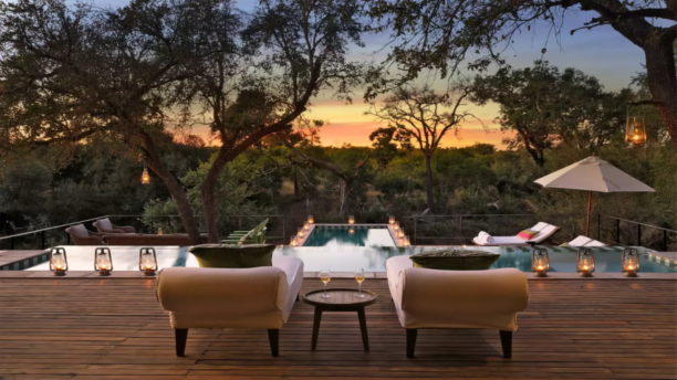 Outdoor seating area with a long infinity edge pool surrounded by lanterns, and with two sun loungers facing the pool and forest beyond