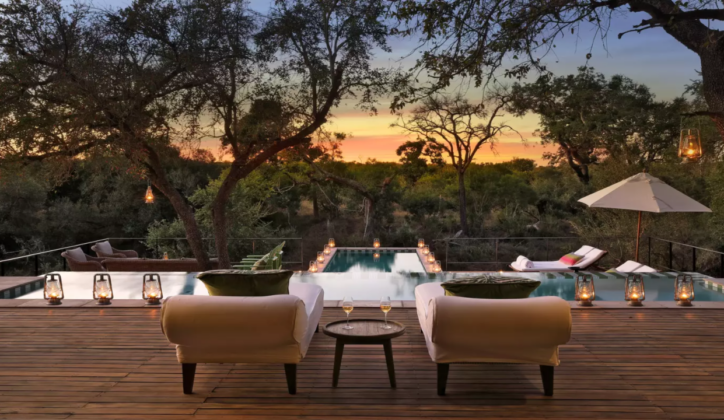 Outdoor seating area with a long infinity edge pool surrounded by lanterns, and with two sun loungers facing the pool and forest beyond