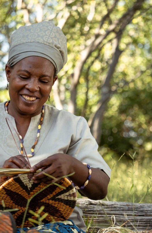 A smiling woman wearing traditional Botswanan clothing and headwrap weaving