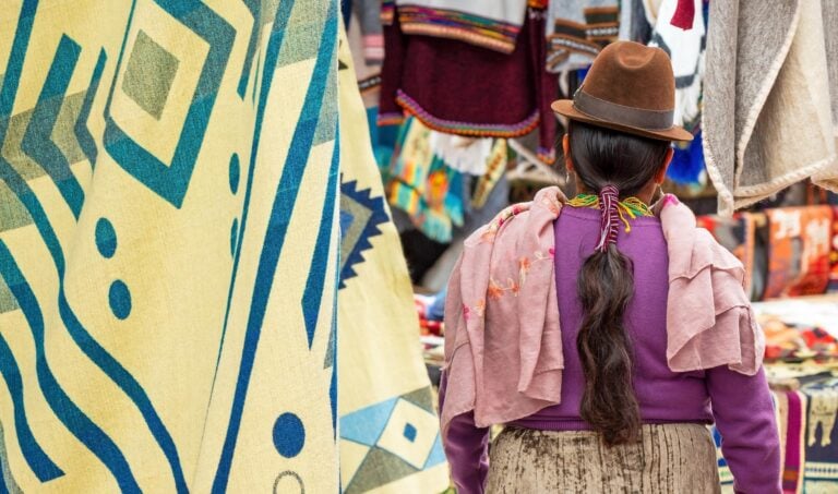 Indigenous Ecuadorian Otavalo woman in traditional clothing, hat and hairstyle on Otavalo local market with textile and fabric stalls, Ecuador.