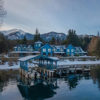 The blue facade of Las Balsas hotel covered in snow, with a lake and boat house in the foreground