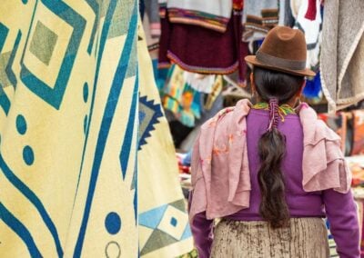 Indigenous Ecuadorian Otavalo woman in traditional clothing, hat and hairstyle on Otavalo local market with textile and fabric stalls, Ecuador.