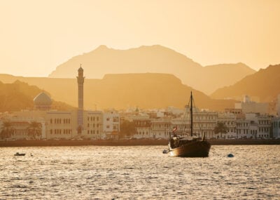 A traditional Dhow floating in the water with Muscat Old Town in the background