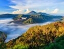 Majestic volcanic landscape with lush greenery, mist rolling through valleys, and distant mountains under a blue sky with fluffy clouds.